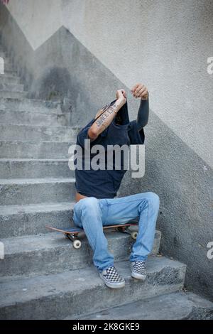Corpo pieno di irriconoscibile hipster maschile che tacca la t-shirt mentre siedi sullo skateboard alle scale durante la pausa Foto Stock