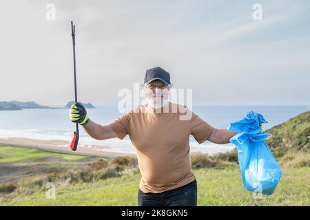 Allegro anziano portatore volontario maschile in guanti protettivi e cappello che tiene in mani sollevate pinze spazzatura e sacco di cestino pieno di rifiuti dopo la raccolta rifiuti Foto Stock