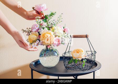 Donna fa bouquet in vaso di fiori rosa giallo arancio sul tavolo a casa. Primo piano di rose inglesi fresche con cestino in metallo. Foto Stock