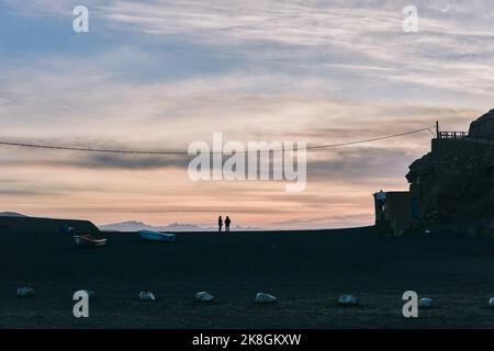 Silhouette di viaggiatori irriconoscibili che si trovano su un'ampia spiaggia di sabbia vicino a una vecchia casa e a una scogliera rocciosa contro il cielo nuvoloso del tramonto durante il viaggio a Fuerteve Foto Stock