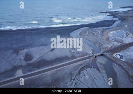 Vista mozzafiato del drone della spiaggia vulcanica nera situata vicino alla strada e alle montagne innevate sulla costa del mare ondeggiante in una serata tempesta in Islanda Foto Stock