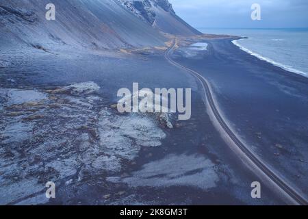 Vista mozzafiato del drone della spiaggia vulcanica nera situata vicino alla strada e alle montagne innevate sulla costa del mare ondeggiante in una serata tempesta in Islanda Foto Stock