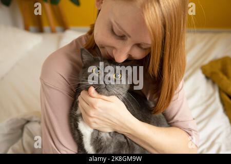 Donna zenzero positiva in pigiama seduta con gli occhi chiusi su comodo letto con gambe incrociate e carezzante teneramente adorabile gatto con zampe bianche Foto Stock