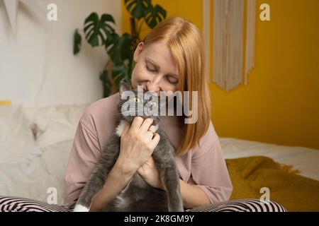 Donna zenzero positiva in pigiama seduta con gli occhi chiusi su comodo letto con gambe incrociate e carezzante teneramente adorabile gatto con zampe bianche Foto Stock