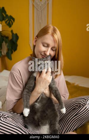 Donna zenzero positiva in pigiama seduta con gli occhi chiusi su comodo letto con gambe incrociate e carezzante teneramente adorabile gatto con zampe bianche Foto Stock