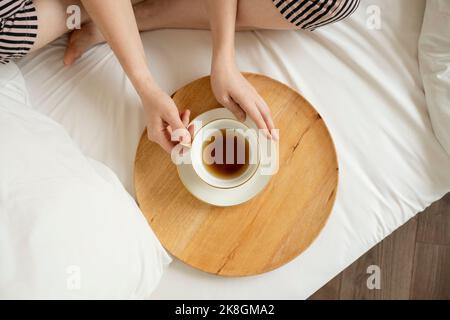 Dall'alto di raccolto irriconoscibile donna a piedi nudi in pantaloni a righe seduta su comodo letto con gambe incrociate e bere tè aromatico servito su woo Foto Stock