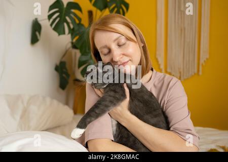 Donna zenzero positivo in pigiama seduta con gli occhi chiusi sul comodo letto carezzando teneramente adorabile gatto con zampe bianche Foto Stock