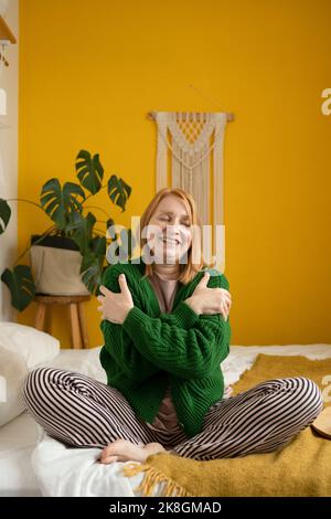 Allegro giovane zenzero donna in verde caldo maglia cardigan e pigiama seduta su comodo letto con gambe incrociate e riscaldarsi in camera da letto con h Foto Stock