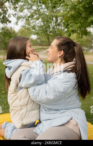 Vista laterale di felice mamma e figlia in outerwear sorridendo e abbracciandosi mentre si siede su coperta gialla il giorno d'autunno nel parco Foto Stock