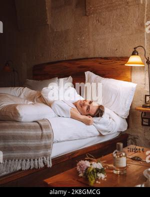 Giovane ragazza turista felice in accappatoio bianco e asciugamano sulla testa sorridendo e guardando la macchina fotografica mentre sdraiato su un comodo letto in camera d'albergo durante il t Foto Stock