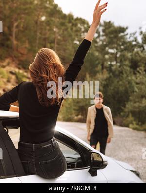 Vista sul retro di una donna irriconoscibile in abiti casual seduti sul finestrino dell'auto che sventolano al ragazzo in campagna Foto Stock