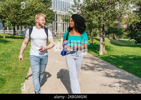 Allegri turisti multirazziali maschi e femmine con zaino e borsa a passeggiare lungo il vicolo nel parco cittadino Foto Stock