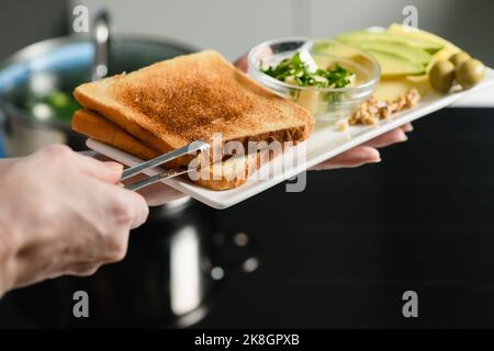 La mano della donna mette pane tostato appena sfornato su un piatto. Avocado a fette, burro, formaggio per un panino sano. Foto Stock