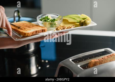 La mano della donna mette pane tostato appena sfornato su un piatto. Avocado a fette, burro, formaggio per un panino sano. Foto Stock