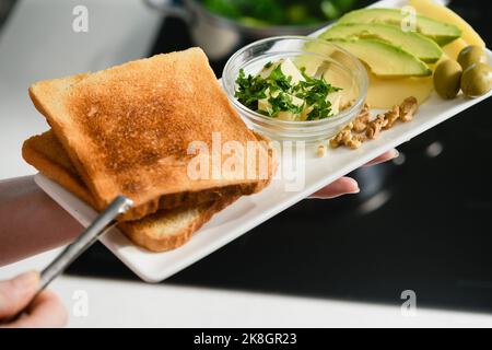 La mano della donna mette pane tostato appena sfornato su un piatto. Avocado a fette, burro, formaggio per un panino sano. Foto Stock