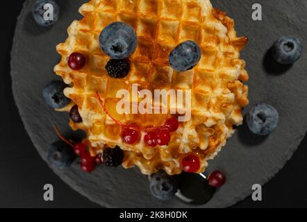 Waffle belgi freschi e croccanti con mirtilli freschi, more e ribes rossi, versati con vista dall'alto sul miele fresco su sfondo nero Foto Stock