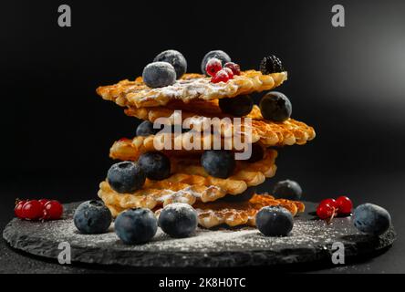 Waffle belgi freschi e croccanti con mirtilli freschi, mirtilli neri e ribes rossi cosparsi di zucchero a velo su un piatto di pietra nera Foto Stock