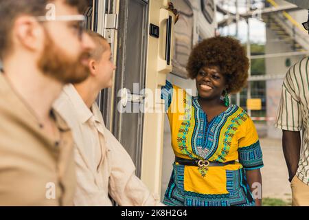 Allegra e colorata donna nera con acconciature afro sorridente ai suoi amici caucasici. Concetto di motorhome. Breve pausa prima di un lungo viaggio verso il nuovo luogo. Foto di alta qualità Foto Stock