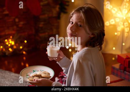 Carino ragazzo pronto per Babbo Natale in attesa con biscotti e latte. Credenza nella fiaba Foto Stock