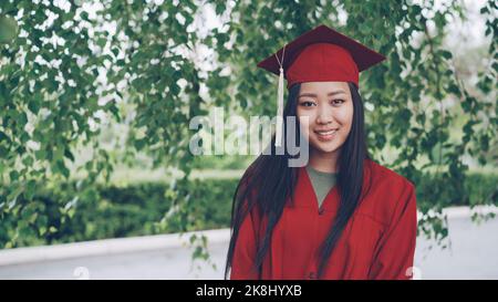 Ritratto di attraente ragazza asiatica laureata studente di successo in abito rosso e Malta-board in piedi sul campus, sorridendo e guardando la fotocamera. Concetto di gioventù e istruzione. Foto Stock