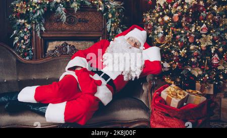 Babbo Natale dorme in una stanza decorata vicino all'albero di Natale con un sacco di regali, l'uomo in costume è sdraiato sul divano. Vacanza e presenta concetto. Foto Stock