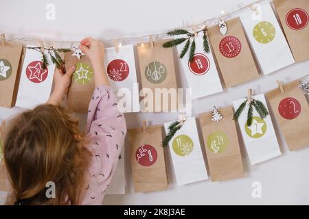 Il bambino del toddler ottiene il nuovo compito di calendario dell'avvento, in attesa di Natale. Umore festivo per la ragazza curly carina. Il bambino prende il sacchetto di avvento con le date dalla parete a. Foto Stock