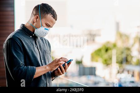 Mettetevi in contatto con i vostri clienti, vogliono sentire da voi. Un giovane uomo d'affari che indossa una maschera e usa uno smartphone sul balcone di un ufficio. Foto Stock