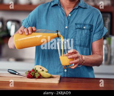 La migliore bevanda a colazione. Una donna anziana irriconoscibile che si versa un bicchiere di succo d'arancia mentre prepara la colazione in cucina. Foto Stock