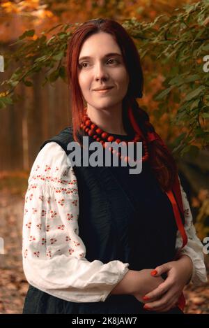 Giovane donna sorridente rossa vestita tradizionali vestiti ricamati ucraini e perle rosse guardando lontano. Vestito vintage. Verticale a metà lunghezza Foto Stock