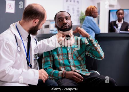 Un uomo afroamericano ferito che indossa un colletto che parla con un medico su appuntamento nella lobby della sala d'attesa. Paziente con dolore con rinforzo cervicale in schiuma dopo lesioni da incidente, in trattamento. Foto Stock