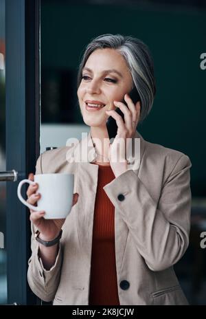 Sigillare gli affari uno dopo l'altro. Una donna d'affari matura attraente che si levano in piedi da sola e che parla sul suo cellulare mentre godendo una tazza di caffè. Foto Stock