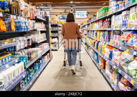 una donna con un carrello cammina tra le file di scaffali in un negozio di alimentari Foto Stock