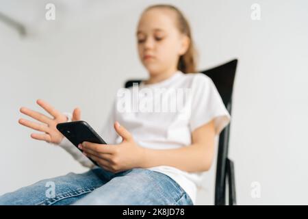 Primo piano basso angolo di vista della bambina bionda con braccio rotto avvolto in cerotto bianco utilizzando la tastiera smartphone seduta sulla sedia in stanza leggera. Foto Stock