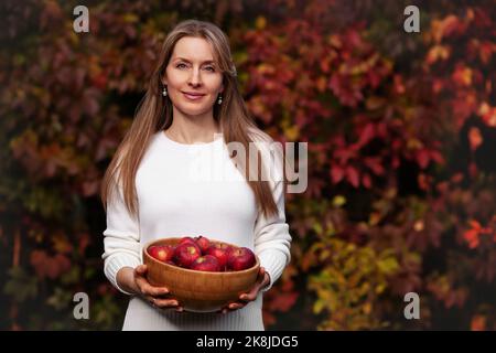 Donna raccolta e tenendo ciotola di mele rosse sullo sfondo delle foglie autunnali Foto Stock