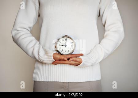 Tempo. Mani femminili con sveglia vintage con le mani sul quadrante. Isolato su sfondo grigio. Ventre di donna in maglione bianco Foto Stock