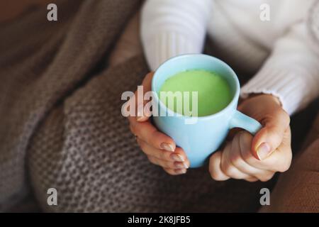 Primo piano delle mani che tengono una tazza blu di tè Matcha. Modello in maglione bianco e confortevole plaid è seduto a casa. Profondità di campo, spazio vuoto sul beig sfocato Foto Stock