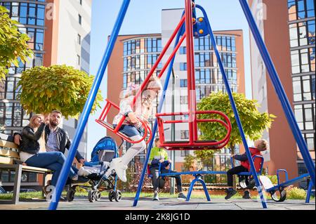 Famiglia felice - padre, madre e bambini che si divertono insieme la mattina di sole. Genitori seduti sulla panchina mentre i bambini giocano nel moderno cortile di edifici residenziali di alta altezza Foto Stock