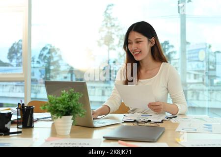 Giovane imprenditrice sorridente che analizza statistiche, grafici finanziari grafici sul suo computer portatile Foto Stock