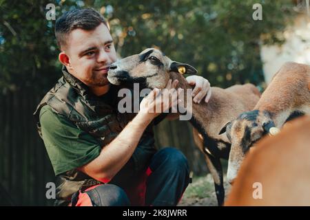 Guardiano con sindrome di Down prendersi cura degli animali in zoo, accarezzando la capra. Concetto di integrazione delle persone con disabilità nella società. Foto Stock