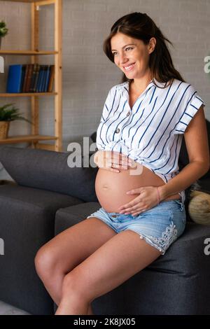 Gravidanza, riposo, persone e concetto di aspettativa. Donna incinta felice che tocca il ventre a casa Foto Stock