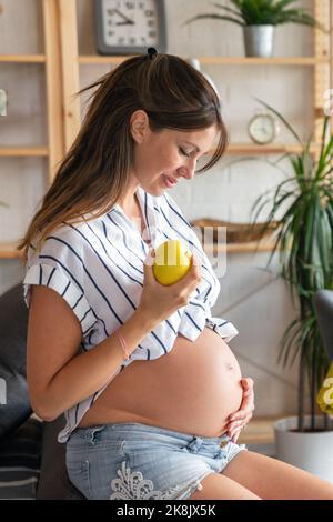 Gravidanza, riposo, persone e concetto di aspettativa. Donna incinta felice che tocca il ventre a casa Foto Stock