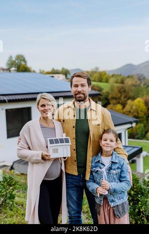 Famiglia felice tenendo modello di carta di casa con pannelli solari. Energia alternativa, risparmio di risorse e concetto di stile di vita sostenibile. Foto Stock