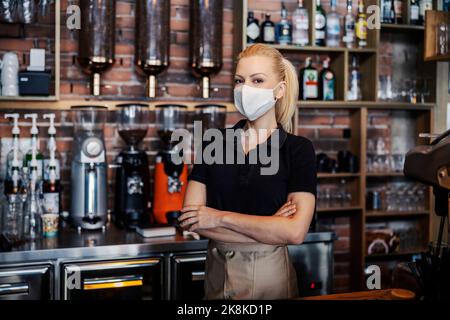 Servizio bevande durante la pandemia del virus corona. La donna si trova al banco del ristorante e indossa una maglietta nera e un grembiule. Una cameriera ha il suo braccio Foto Stock