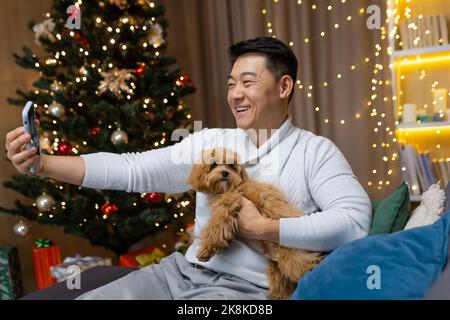 La vigilia di Natale uomo asiatico con un piccolo cane da compagnia che parla in videochiamata con gli amici, uomo seduto sul divano vicino all'albero di Natale festeggia il nuovo anno in attesa di amici di partito. Foto Stock