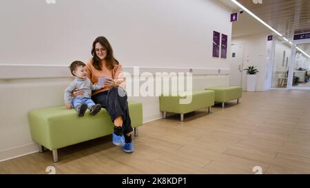 Madre e bambino con un telefono cellulare in clinica in attesa di un pediatra. Genitore con un bambino con uno smartphone nelle mani. Il bambino è un ragazzo Foto Stock