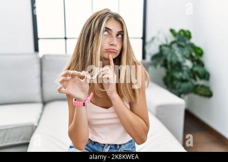 Giovane donna bionda che tiene invisibile aligner ortodontico pensiero concentrato sul dubbio con un dito sul mento e guardando in su chiedendo Foto Stock