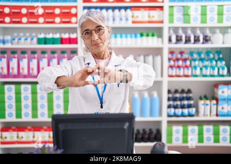 Donna di mezza età con tatuaggi che lavorano in farmacia sorridendo in amore facendo forma di simbolo del cuore con le mani. Concetto romantico. Foto Stock