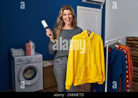 Giovane donna che usa il rotolo di lanugine per rimuovere i capelli dai vestiti che attaccano la lingua fuori felice con l'espressione divertente. Foto Stock