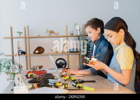 preteen ragazzo utilizzando laptop vicino a una ragazza che tiene parte meccanica del modello robotico, immagine stock Foto Stock