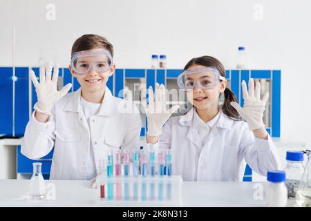 sorridendo i bambini con camici e occhiali bianchi che mostrano le mani nei guanti in lattice vicino alle provette in laboratorio, immagine di scorta Foto Stock